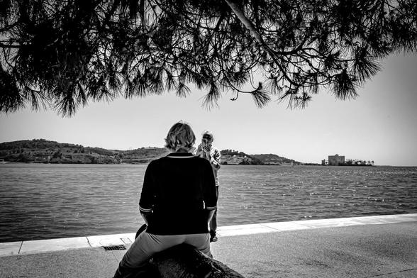 Two people near a waterfront, with one sitting on a log facing away and the other standing taking a photo. Overhead tree branches create a natural frame. A calm body of water and a distant shoreline with hills are visible in the background, along with a large building on the right. The image is in black and white.