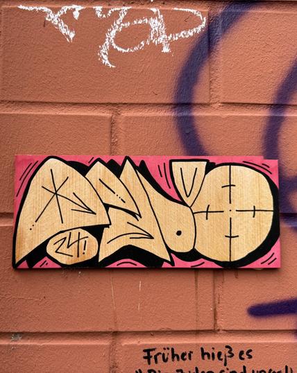 Pinke Kachel mit gelb-schwarzer Bemalung auf rotem Backstein. Motiv: Kreise und andere geometrische Figuren auf denen Pfeile sind.