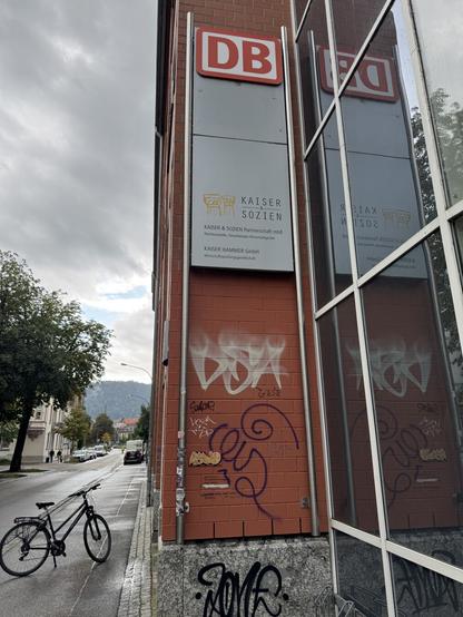 Gebäude mit Schildern (DB-Logo, Anwaltskanzlei), daneben eine Straße und ein Fahrrad. Klein ist die Kachel zu sehen.