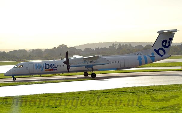 Side view of a high-winged, twin propellor-engined airliner taxiing from right to left.
The plane is mostly white, with a blue belly, and two-tone blue "FlyBe" titles on the forward fuselage, tail, and the sides of the engine pods under the wings.
Green grass fills the foreground, next to a taxiway which is reflecting bright sunlight.
Trees line the airfield perimeter in the background, running from one side of the image to the other.
Rolling hills in the distance vanish into the bright  but hazy sky.