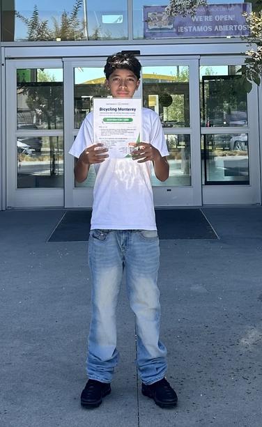 A teen is standing in front of a Salinas library, holding a Bicycling Monterey poster.