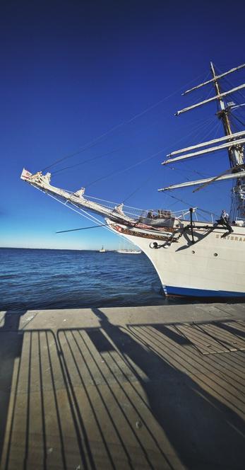 A low-angle, vertical shot of the bow and part of the main body of a large, white sailing ship docked at a pier. A part of ship's name "STATSRAA" is visible on the bow. The water is a deep blue and choppy, and the sky is a clear, bright blue. The sun casts long shadows of a railing onto the pier in the foreground. In the distance, other ships can be seen on the horizon.