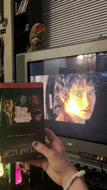 Me holding up a VHS box of 
“Lord of the Rings The Fellowship of The Ring” exclusive behind the scenes features
In the background the tape is playing on a Sony CRT with Elijah Wood looking at the viewer