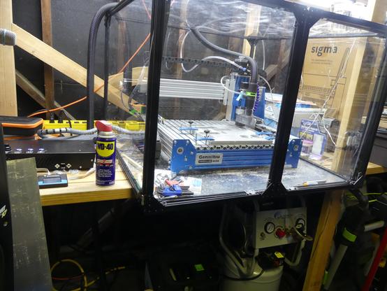 A workspace with a CNC machine enclosed in a transparent protective casing. The machine is blue and silver, with a label reading "Genmitsu." It is mounted on a workbench, surrounded by various tools and equipment. On the left side of the image, there is a can of WD-40 lubricant, a black electronic device, and a digital display. The background includes wooden beams and a cardboard box with the word "sigma" visible. The workspace appears cluttered with tools and cables, indicating an active work environment.