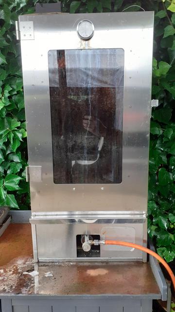 Ein rechteckiger Edelstahl-Räucherschrank mit Glasscheibe in der Tür (ca 40cm x 80cm) steht auf einem Garten-Holzschrank vor einer mit Efeu berankten Palisadenzaun (den man jedoch nicht mehr sehen kann).