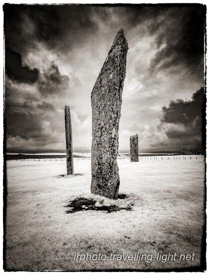 Toned black and white infrared photo of three large flat standing stones in a field, with a central stone and two others behind, all under a menacing sky.