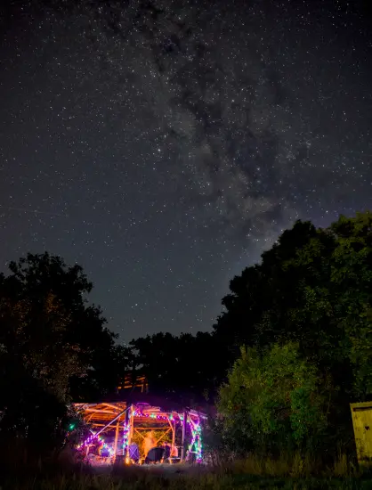 bild im hochformat bei nacht, im unterem dritten unter bäumen eine kleine beleuchtetet holzhütte, in der ein lagerfeuer brennt, drüber der sternhimmel mit der milchstraße