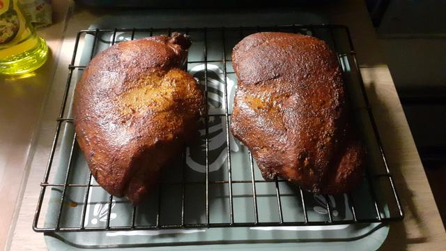 Zwei dunkelbraune marinierte und geräucherte Putenbrüste liegen nebeneinander auf dem Räuchergitter auf einem grauen quadratischen Tablett zum abkühlen. Das Tablett liegt auf einer Buche farbenen Küchenarbeitsfläche.