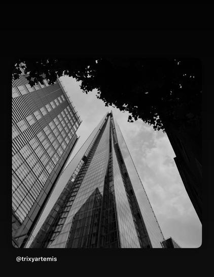 Black and white photograph taken from the base of The Shard, looking upward; a tree occupies the top right corner of the frame, while a second glass building rises to the left, creating a dramatic contrast of natural and architectural forms.