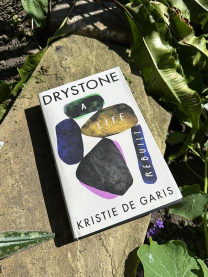 A picture of the book Drystone A Life Rebuilt by Kristie De Garis. The book is white with stones arranged on the front. It is sitting on a small stone wall next to my pond.