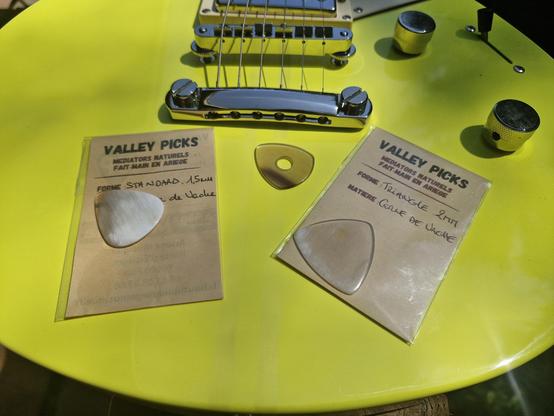 Trois médiators en corne de vache sont posés sur la table jaune flashy d'une guitare électrique. Deux sont encore emballés dans leur petite enveloppe transparente avec un papier gris où son écrits au stylo leur forme ("triangle", "standard") et leur matière ("corne de vache").
