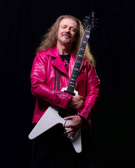 Vito C. mit pinker Lederjacke vor schwarzem Hintergrund umarmt seine weiße Gibson Flying-V Gitarre