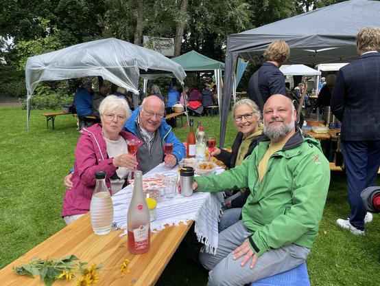 4 Menschen am Picknicktisch prosten der Kamera zu.
