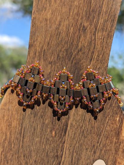 A close-up of a Moroccan Tiles bracelet I made using tila beads, drop beads, and seed beads. The tilas are a metallic reddish bronze with a hint of a blue sheen, the drops are amber yellow with an AB (iridescent) finish, and the seed beads are a raspberry-esque pink; also with an AB finish. The bracelet is displayed in direct sunlight and modeled on a flat wooden hand mannequin. You can vaguely make out a blue sky and green trees in the background.