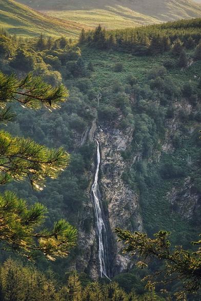 A long shot of a tall, narrow waterfall cascading down a sheer rock face covered in lush green foliage. The photo is taken from a high vantage point, looking down into a valley. Pine branches with soft, glowing sunlight on them frame the top and bottom of the image. The valley floor is covered in trees and the hills beyond are a mix of green grass and trees.