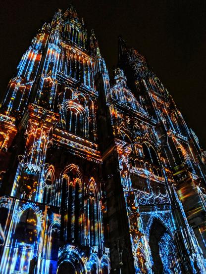 A dramatic, low-angle shot of a magnificent Gothic cathedral at night, its elaborate facade illuminated by a vibrant, multi-coloured light show. The light patterns, in shades of blue, orange, and white, highlight the intricate architectural details of the two towering spires and ornate arches. The contrasting light and dark creates a powerful and almost ethereal effect.