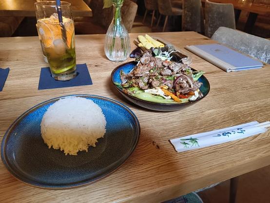Das Bild zeigt ein Essen auf einem Holztisch in einem Restaurant. Auf der linken Seite befindet sich ein blauer Teller mit einem Haufen weißer Reis. Rechts daneben ist ein weiterer blauer Teller mit einem Gericht, das aus geröstetem Rindfleisch, Gemüse und Salat besteht. Die Fleischstücke sind in Streifen geschnitten und mit Gewürzen und Kräutern garniert. Neben dem Gericht liegen ein paar gefaltete Servietten und ein Paar Essstäbchen in einer weißen Hülle mit grünen Zeichnungen. Im Hintergrund ist ein Glas mit einem Getränk, das mit Eiswürfeln und Zitronenscheiben garniert ist, sowie ein kleines, transparentes Vase mit Wasser und Blumen zu sehen. Ein Notizbuch liegt auf dem Tisch, und die gesamte Szene wirkt einladend und gemütlich.