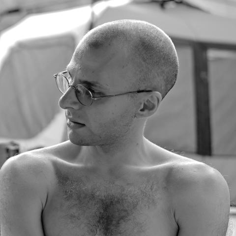 My camp neighbor for 4 days, Matt. The black-and-white photo shows a white man in his 20s or early 30s, head shaved, shirtless, from the chest up. He is looking to his right (the left of the viewer), his round Gandhi-style glasses reflecting the sky.