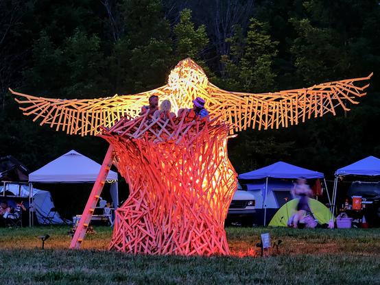 Three figures sit about 3m (15 feet) above a grassy field in an oddly-shaped tower made of wooden slats. The tower is a bird's nest. created to be burned at a regional burn (POrtal burn in NY state). Above and behind the nest is a large wooden bird of the same construction, wings spread approximately 6-7m (20 feet, approximately). The piece is lit with rose, tangerine, and golden light. Below and behind the bird and nest are some tents and trees.