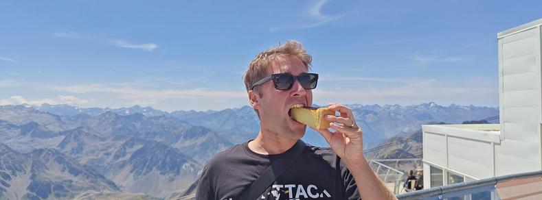 Photo de moi croquant une part de flan sur la terrasse du Pic du Midi, avec un panorama de montagnes dans mon dos.