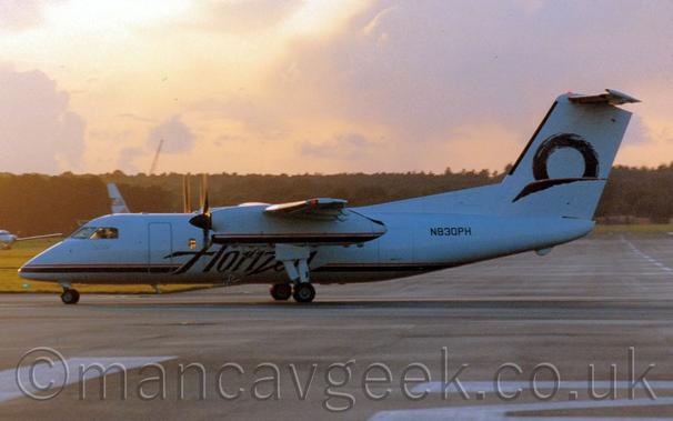 Heavily backlit side view of a high-winged, twin propellor-engined airliner parked facing to the left.
The plane is almost entirely white, except for the black and red stripe running along the bottom of the fuselage, overlaid with black "Horizon" titles under the wings, and the partial black circle over a black brush stroke on the tail.
A grey taxiway fills the foreground, extending off into the distance in the background on the right of the frame, with grass in the background on the left.
Trees in the distance meet the orange sky, with purple-grey clouds, and a bright spot in the top left corner that is the sun which looks as if it will soon be setting.