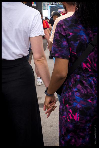 Two people holding hands in a crowded street, one wearing a white t-shirt and dark trousers, the other in a colourful purple and pink patterned dress, at the Cork Pride after-party on Kennedy Quay.