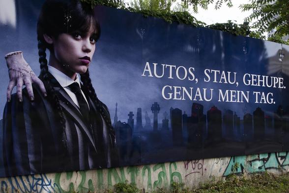 farbfotografie, querformat. schwarzblaue werbebande mit der aufschrift: autos, stau, gehupe. genau mein tag. links daneben eine figur mit einer einzelnen genähten hand auf der schulter.

Color photograph, landscape format. Black-blue advertising banner with the inscription: Cars, traffic jams, honking horns. Just my kind of day. To the left, a figure with a single sewn hand on the shoulder.