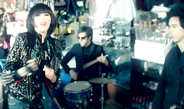 Screenshot of the band from the Zero video. Karen O has kind of a Pat Benatar thing going on with a black bob haircut and rhinestone encrusted leather jacket. Nick and Brian look like they just stepped out the matrix 9 years late, with completely black outfits with sunglasses, and give more of a school shooter vibe than massively talented rockstar.