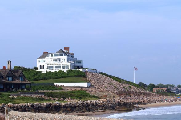 A large white multi-story house with multiple chimneys sits on top of a grassy hill overlooking a rocky coastline and sandy beach. The hill is reinforced with stone riprap, and a tall flagpole with an American flag is positioned on the slope near the right side of the image. Other smaller houses with dark roofs and shingles are visible nearby. The beach below the hill has waves reaching the shore and a group of people gathered along the sand. The sky is clear with light blue color, and the ocean is calm.