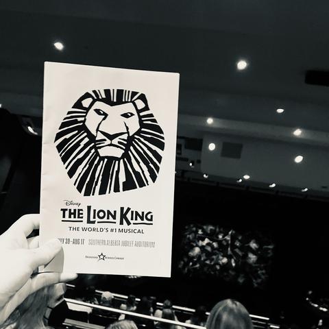 A black and white photo of a child’s hand holding a performance program for the Lion King. People and the stage are in the background.