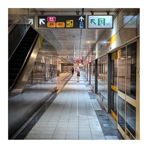 This image shows a clean and well-lit underground metro or subway station platform. The platform is tiled with light-colored square tiles, and the ceiling has evenly spaced lights. On the right side, there are glass doors with yellow horizontal lines that separate the platform from the train tracks, indicating a platform screen door system for safety.

On the left side of the image, there is a metal escalator going upwards, with its dark steps and shiny metal sides reflecting some light. Above the escalator, there is a large sign with multiple directional indicators. The signs include Chinese characters and English words like "Exit," along with arrows pointing upward and to the left. The sign also shows station names such as "Tamsui," "Huilong," and "Luzhou," suggesting this is a metro station in a Chinese-speaking city, likely Taipei.

Two people are visible walking along the platform in the distance. One is closer to the center of the platform, facing to the right, and another is further back, walking away from the camera. The overall atmosphere is calm and orderly, with no crowd visible. The station appears modern, clean, and spacious.