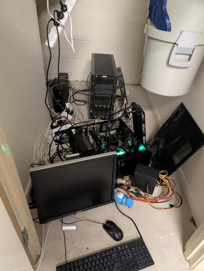 a total mess, a pile of computer on a concrete floor. 

from top to bottom we have a dell poweredge t420 workstation, leaning against it is a rack mount switch. To the left of those is a UPS, then in front of that is a PC with the side off, laying horizontally, with a 360mm radiator just hanging out of it. There's a disconnected power supply and a can of compressed air in front of that, with a monitor in front of all of it and a keyboard/mouse.

Also you can see the powerboard hanging from the top of frame and a central vacuum system attached to the wall on the left.

there is also just cables running between everything.