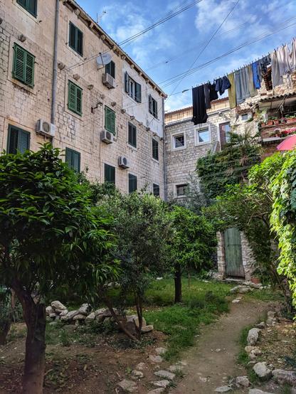A small, enclosed courtyard between stone buildings, with a dirt path leading to a rustic green door. Lush green trees and bushes fill the space, and there are clothes hanging on a washing line strung between the buildings above. The stone walls of the buildings have multiple windows with green shutters, and there are several air conditioning units visible. The scene has a rustic, lived-in feel, suggesting a Mediterranean or Southern European location.