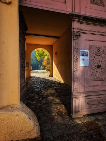 A sunlit archway opens to a cobbled courtyard. The large, carved wooden door on the right, painted a faded pink, is ajar, revealing the entrance to a building. Sunlight streams through the arch, illuminating the wet cobblestones and casting a long shadow from the doorway. In the distance, an archway opens to an outdoor area with green and yellow trees, suggesting an autumnal scene. A poster is pinned to the pink door, and the yellow-painted wall of the archway is bathed in warm light.
