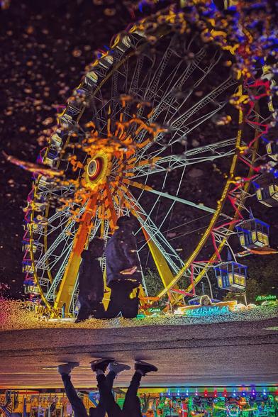 Das Bild zeigt die nächtliche Pfützenspiegelung eines Riesenrads, das in den Farben Gelb, Orange Rot und Blau strahlt. Im Vordergrund gehen zwei Personen, deren Beine allerdings nicht in der Spiegelung zu sehen sind, sondern im Originalbild, das in diesem Fall unten zu sehen ist, weil das Bild um 180 Grad gedreht wurde.
