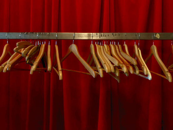A horizontal metal bar seen in the top half of the frame supporting a number of wooden clothes hangers. The background shows heavy deep red curtains, the kind often seen in cinemas and theatres.