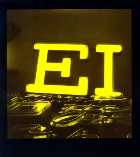 Das Polaroid zeigt die beiden Großbuchstaben "EI" als Teil einer großen Leuchtreklame. Die Buchstaben leuchten in einem extrem hellen, fast neongelben Farbton, der die gesamte Szene dominiert und in ein monochromes Licht taucht.

Im Vordergrund, unterhalb der leuchtenden Buchstaben, befindet sich eine unordentliche Ansammlung von vielen weiteren, ausgeschalteten Neonröhren Buchstaben.