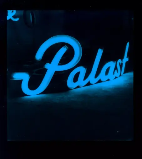 Das Polaroid zeigt eine leuchtende Neonreklame mit dem Wort "Palast" in einer geschwungenen, kursiven Schrift. Die Röhren der Reklame strahlen in einem intensiven, hellen Blauton (Monochrome Blau vom Polaroid)
