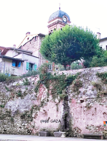 a medieval wall covered in plant growth bears the call 'free gaza'. above is a verdant, full, wide tree behindwhich are houses & the clocktower of a church.