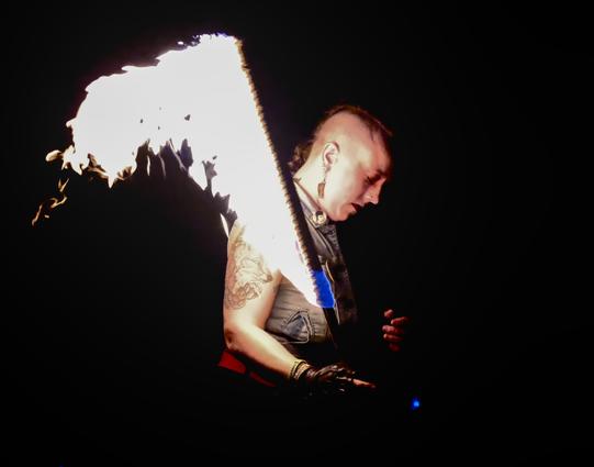 A woman with a shaved/mohawk hairdo, dangly earrings, denim vest, red belt, and fingerless leather gloves holds a flaming sword. Literally flaming. She was spinning it at the time. She is looking downward with her eyes closed but don't be fooled; if you make a move for the fruit from that tree, she will fuck you up.