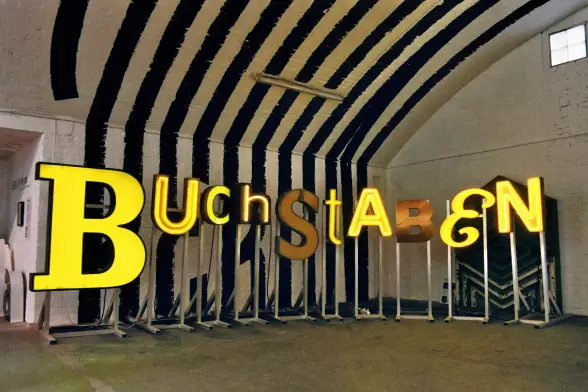 Das Bild zeigt eine Kunstinstallation in einem großen Raum mit einer markanten, gewölbten Decke, die mit breiten, schwarzen Streifen bemalt ist.

Im Zentrum stehen mehrere große, gelbe freistehende Buchstaben, die zusammen das Wort "BUCHSTABEN" bilden. Die Buchstaben sind in unterschiedlichen Stilen und Größen.
