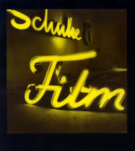 Polaroid zeigt eine Ansammlung von Neonreklamen in einem dunklen Raum, der vollständig in ein intensives, gelbes Licht getaucht ist.

Im Vordergrund und als zentrales Motiv dominiert der leuchtende Schriftzug "Film". Er ist in einer eleganten, geschwungenen Kursivschrift gestaltet.

Dahinter, an der Wand im Hintergrund, ist ein weiterer, etwas kleinerer Neonschriftzug mit dem Wort "Schuhe" zu erkennen.

Die monochrome gelbe Farbgebung stammt vom Polaroid