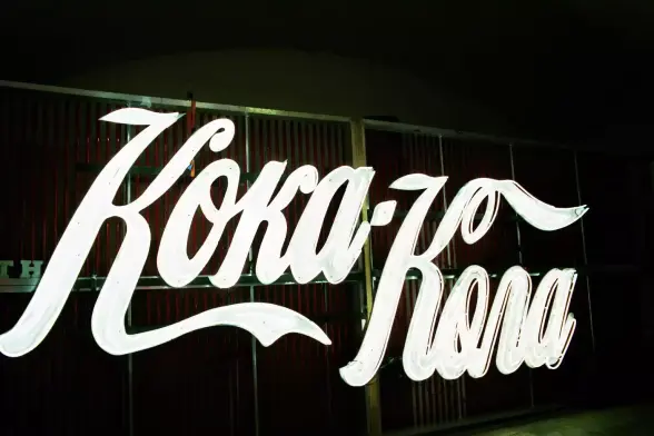 Das Bild zeigt eine große Neonreklame, die den Schriftzug "Koka-Kola" in einer sehr hell leuchtenden, weißen Farbe darstellt. Die geschwungene, dynamische Kursivschrift stammt von der Volksbühne aus dem Stück "Brüder Karamazov".