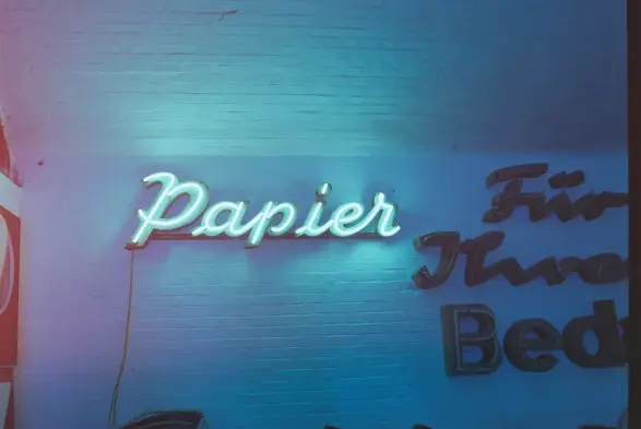 Das Foto zeigt eine leuchtende Neonreklame mit dem Wort "Papier" an einer weiß gestrichenen Backsteinwand.

Der Schriftzug ist in einer geschwungenen, kursiven Schriftart gestaltet und leuchtet hell, während er einen intensiven türkisfarbenen Schein auf die umgebende Wand wirft. (Revolog Color Effekt vom Analogfilm)