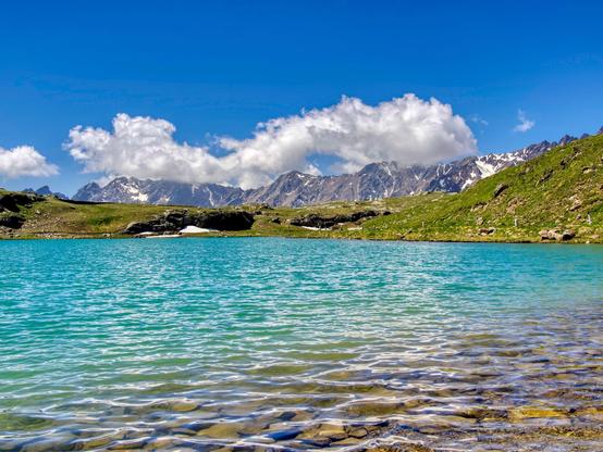 Crystal clear waters of a mountain lake in a sunny and beautiful day. Underwater pebbles con be seen in the foreground, then the water color becomes turquoise, till the green grass of the banks in the background. Mountain peaks, some snowcapped, in the far background.