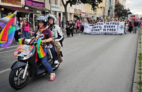 Das Foto zeigt zwei Personen auf einem Motorroller, die der Demo mit dem Fronttransparent vorausfahren. Der Text auf dem Transparen lautet: "No Dyke* is free until evey Dyke* is free - Community Dyke March Berlin 2025"