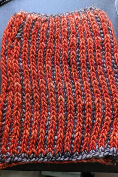 A hand-knit cowl atop a black table. The cowl is made of two colors of yarn: an orangey red and a silvery gray. Both colors are variegated and get lighter and darker.
