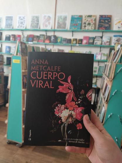 Fotografía con la librería de fondo donde aparece en primer plano el libro: fondo negro, hombre y título a la izquierda, y centrado a la derecha un rampo de flores con insectos que parecen sacados de una pintura. El juego de luces y sombras hace que haya flores que estén ocultas mientras hay otras que resaltan.