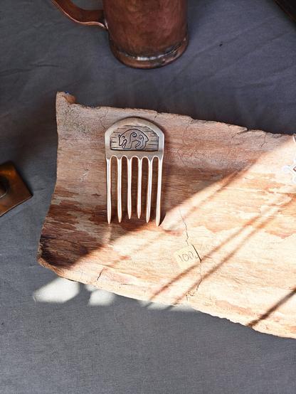 Picture of a table that has objects made of bronze that are for sale. In the middle there is a hair comb that can be used to pull up the hair. The comb has bear decoration carved on it. It lays on top of some piece of wood and sun shines to the wood in triangle shape.