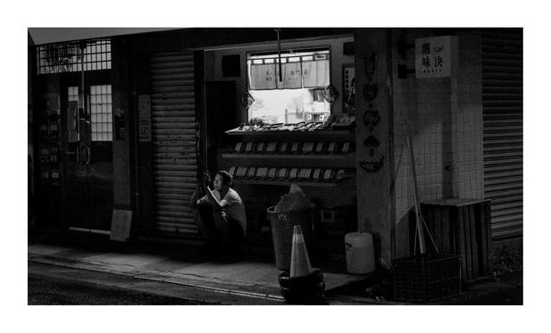 This is a black-and-white photo of a quiet nighttime street scene, likely in Taipei. The photo has a moody, cinematic atmosphere, with strong contrasts of light and shadow.

In the center of the image is a small food stall or shop. The upper half of the shop is brightly lit from inside, illuminating a counter with packaged goods or snacks neatly arranged in rows. Above the counter hangs a short curtain with Chinese writing, suggesting the name and type of business—possibly a snack or specialty shop.

In front of the shop, in the dim light of the street, a person is sitting on the ground, hunched slightly, looking at a smartphone. The glow of the screen faintly lights their face. They appear to be taking a break or waiting, and they seem relaxed but slightly weary.

To the right, a traffic cone and a large trash bin are placed near the sidewalk. The surroundings are framed by closed shutters, tiled walls, and some faded posters or wall graphics. The street is quiet, empty of traffic or people except for the lone figure. The image captures a moment of solitude, everyday life, and calm urban stillness.